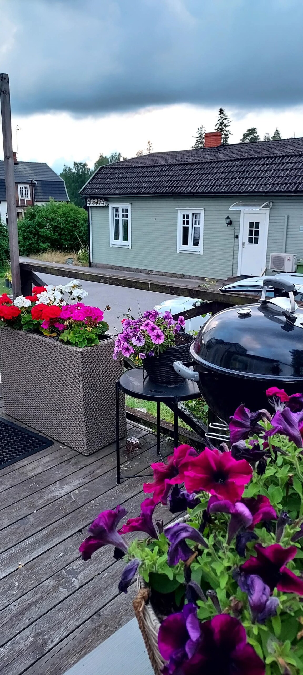 A large deck area is adorned with vibrant flower pots containing colorful blooms, including purple and pink petunias. A charcoal grill is positioned on a small table, inviting outdoor cooking. The house in the background features multiple windows and a traditional roof.