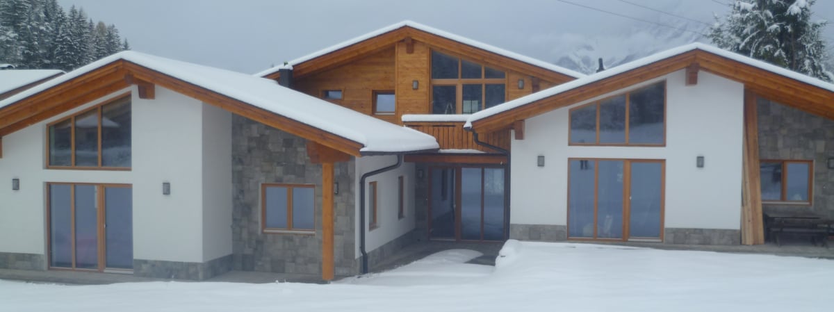 A modern chalet is set against a snowy landscape, characterized by a combination of stone and wood facades. Large windows allow light to enter, while an inviting entrance is framed by a covered porch. The surrounding snow enhances the serene environment.