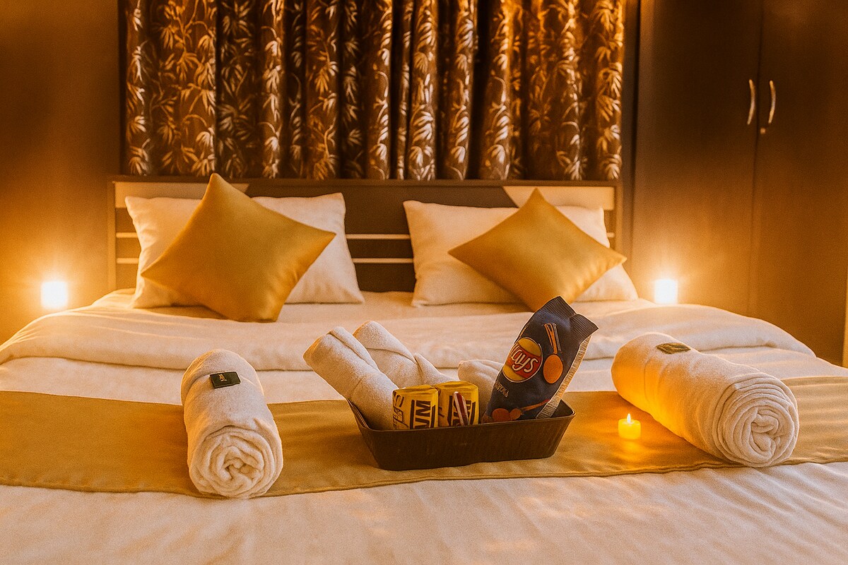 A well-arranged sleeping area features a large bed adorned with gold accents, complemented by decorative pillows. Rolled towels are neatly placed on the bedding, and a tray holds snacks and candles, contributing to a calming atmosphere. The background is decorated with patterned curtains.