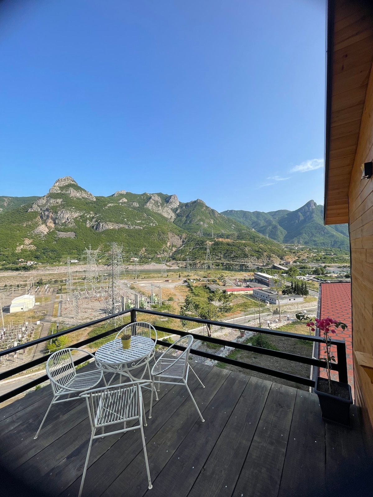 A balcony features a small round table with two chairs, offering unobstructed views of surrounding mountains and valley landscapes. The wooden railing provides a safety barrier while allowing guests to appreciate the natural scenery and expansive sky.