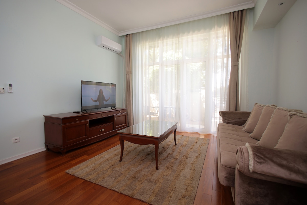 A bright living area features large windows with sheer curtains, allowing natural light to fill the space. A comfortable sofa and a wooden coffee table are arranged on a light-colored rug, while a television is positioned on a wooden cabinet, enhancing the room's inviting ambiance.