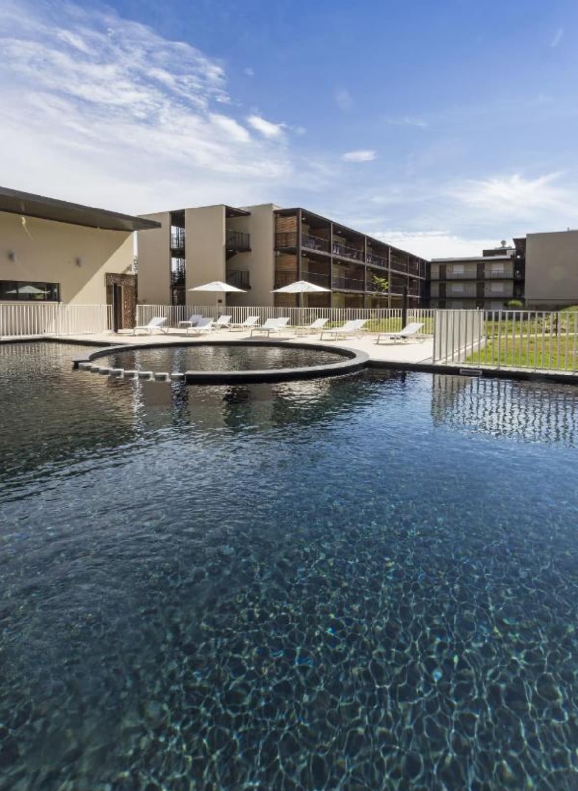A serene swimming pool is depicted, with clear water reflecting the blue sky. Sun loungers are arranged around the pool, accompanied by umbrellas for shade. The modern building in the background features multiple balconies, enhancing the inviting atmosphere of the outdoor space.