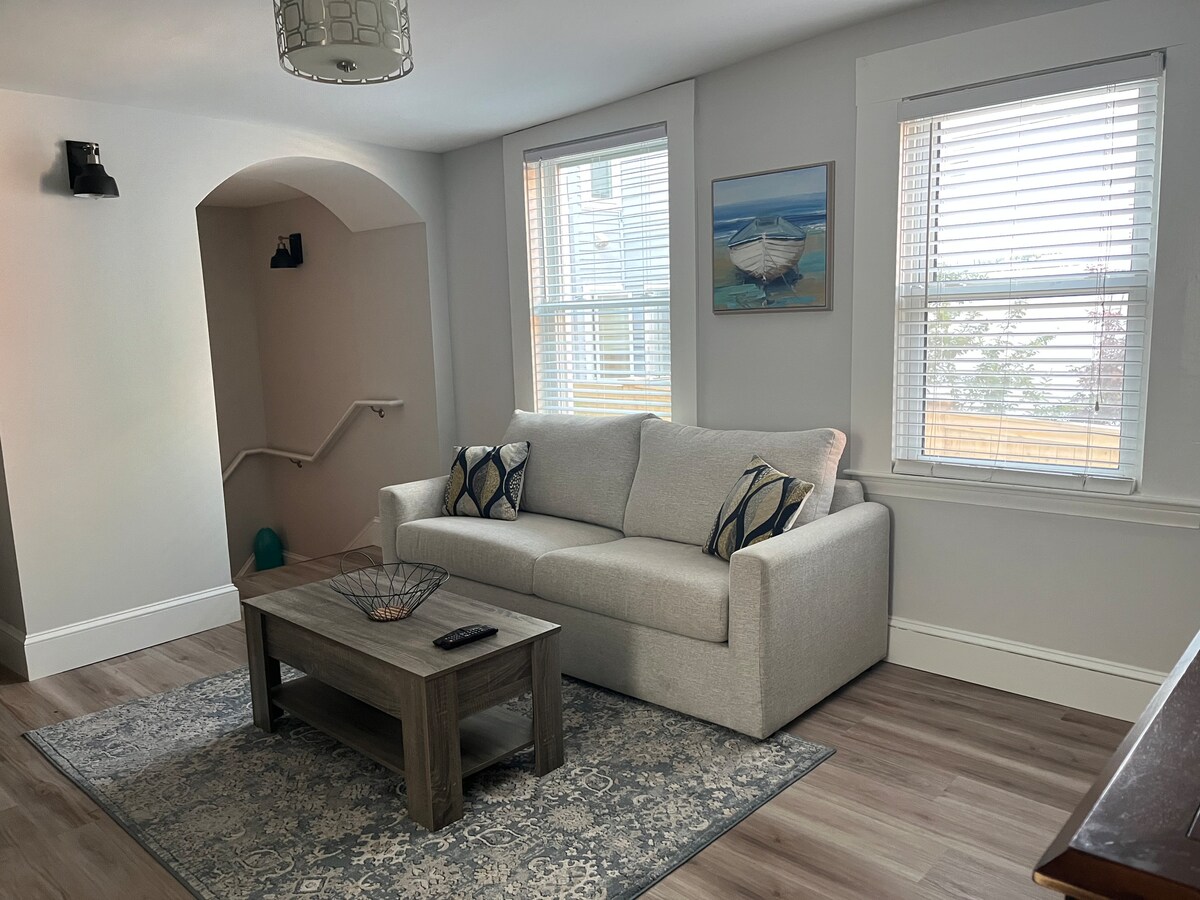 A comfortable living area features a light-colored sofa adorned with decorative pillows. A wooden coffee table is positioned on a patterned area rug. Sunlight filters in through two windows, illuminating the space and highlighting a wall art piece.