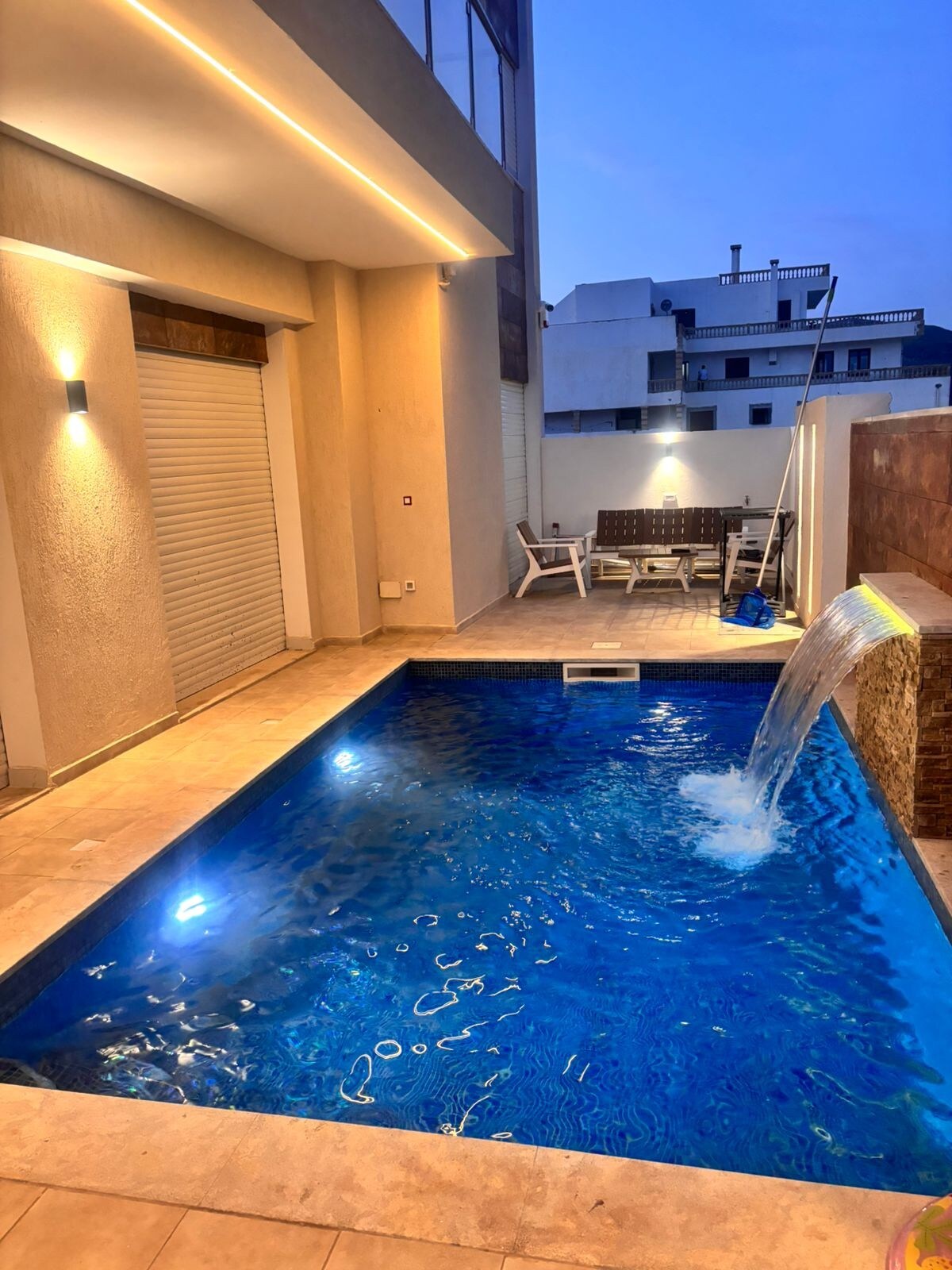An outdoor area features a private pool with a waterfall and illuminated water, creating a calming ambiance. Comfortable seating is present nearby, and the surrounding walls provide privacy. The soft lighting enhances the overall appeal, inviting relaxation in the evening hours.