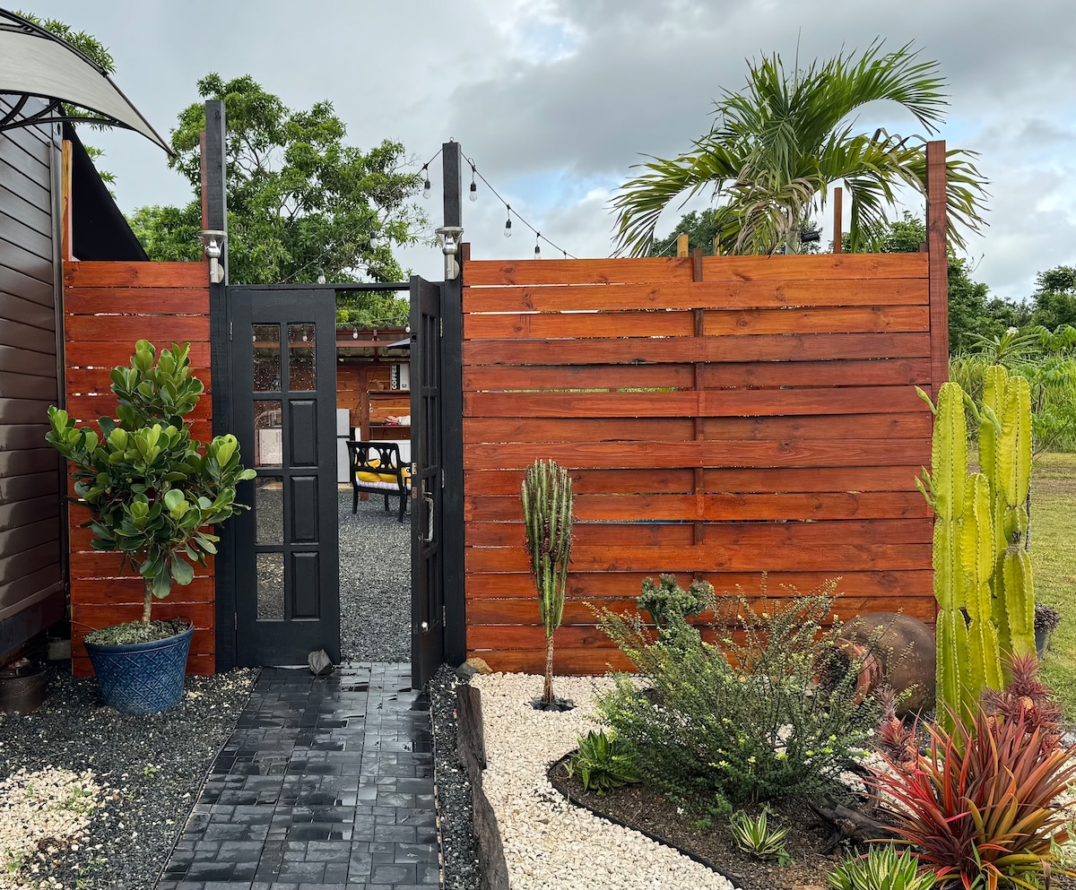 A welcoming entrance is framed by a dark wooden gate, offering a glimpse into a private outdoor space. Lush greenery and decorative plants, including cacti, line the pathway, which is bordered by light gravel and dark stones, creating a serene atmosphere.
