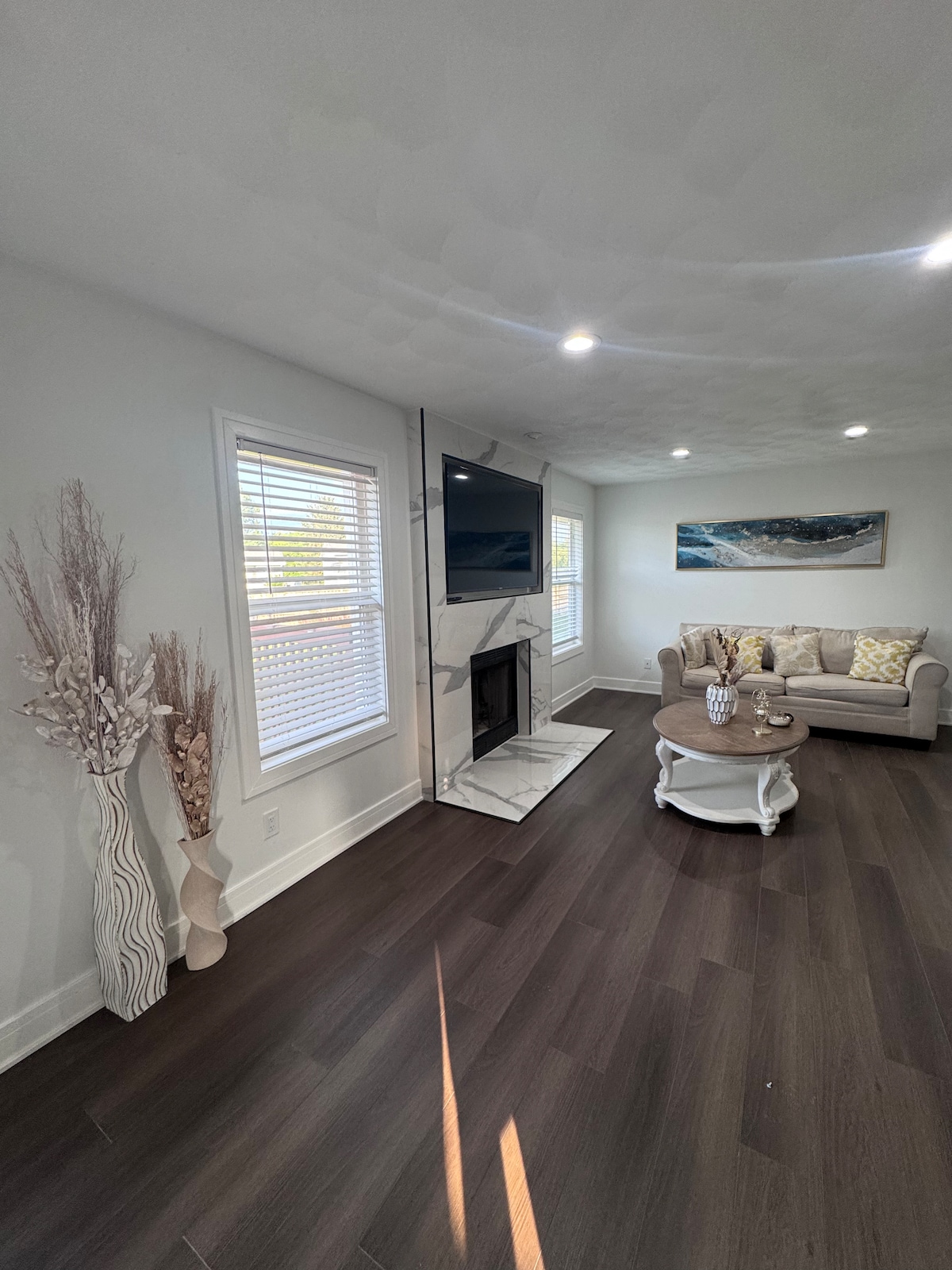 A cozy living area features a light-colored sofa accompanied by decorative cushions. A marble fireplace is positioned beneath a wall-mounted television. Natural light enters through multiple windows, complemented by simple decorative elements like vases and a round coffee table.