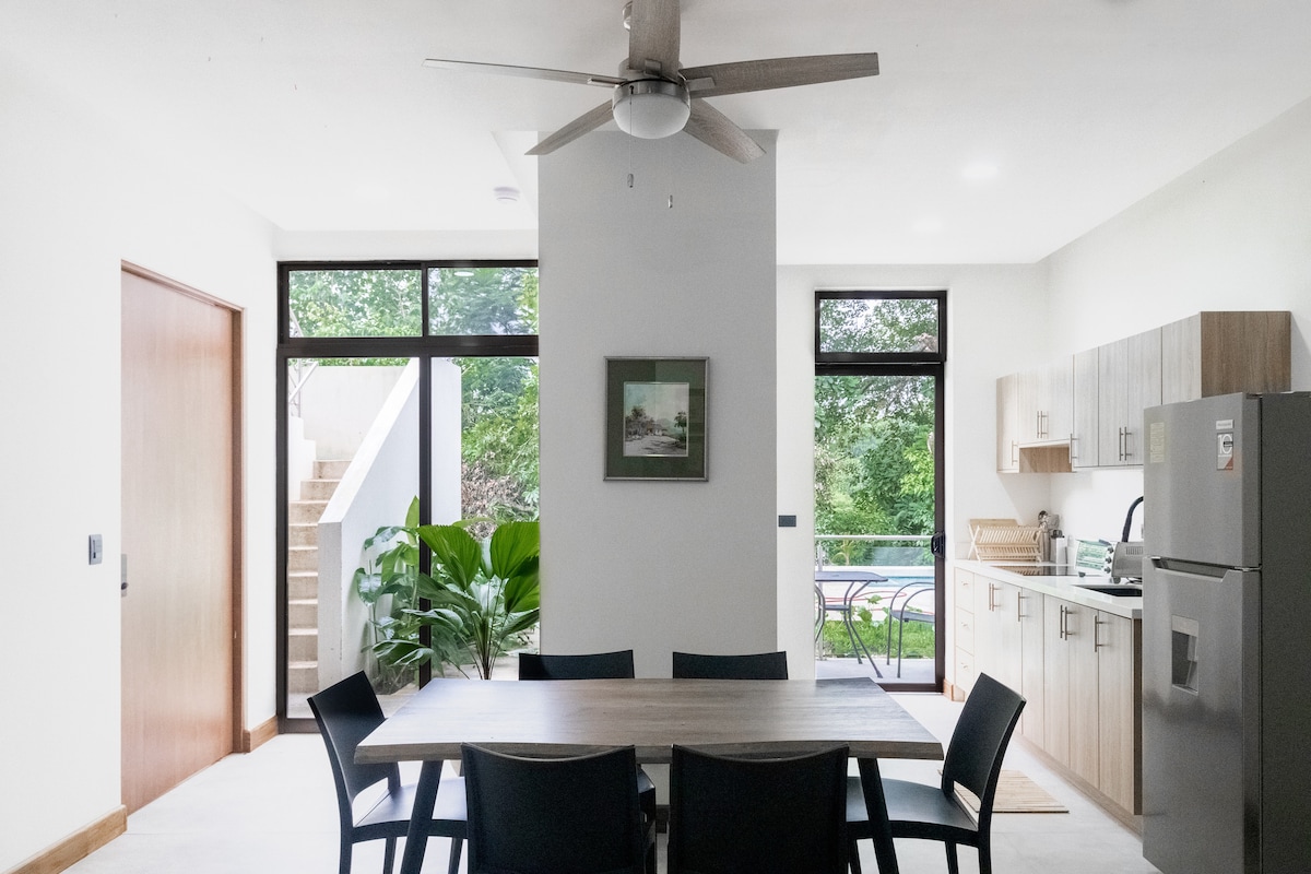 A modern dining area is positioned adjacent to a well-equipped kitchen, featuring a large table surrounded by six chairs. A ceiling fan hangs above, while large windows allow natural light to fill the space. A staircase is visible, leading to additional areas of the apartment.
