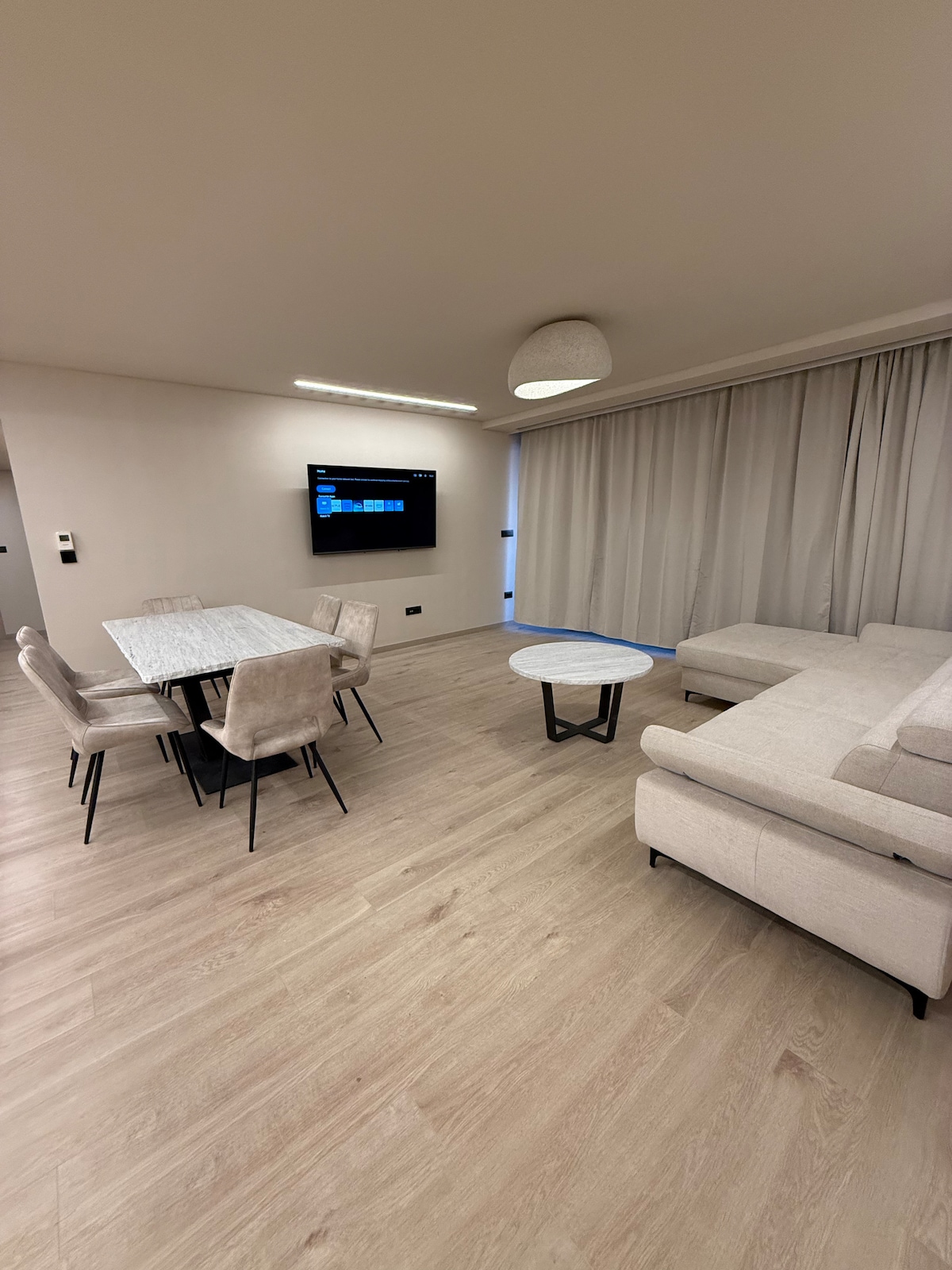 A modern living space features a spacious sectional sofa, a small coffee table, and a dining table with six upholstered chairs. Soft lighting illuminates the area, while large curtains frame a window, offering a sense of privacy and comfort. A wall-mounted television is visible.