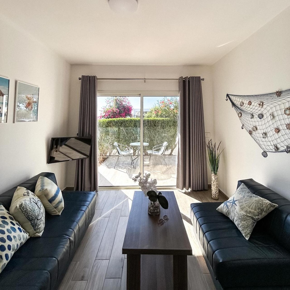 A bright living area is furnished with two dark sofas adorned with patterned cushions. A wooden table sits at the center, and natural light floods in through sliding glass doors that open to a private terrace with outdoor seating. Greenery is visible outside.
