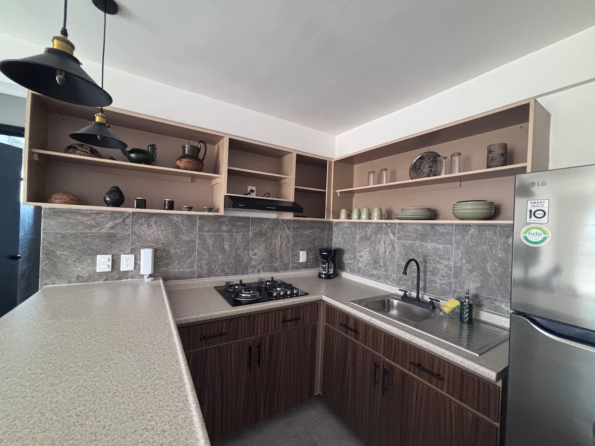 The kitchen is equipped with modern appliances and ample countertop space. Open shelving displays pottery and decorative items. A gas stove and sink are easily accessible, and a coffee maker is positioned on the countertop. The overall layout promotes efficiency and functionality.