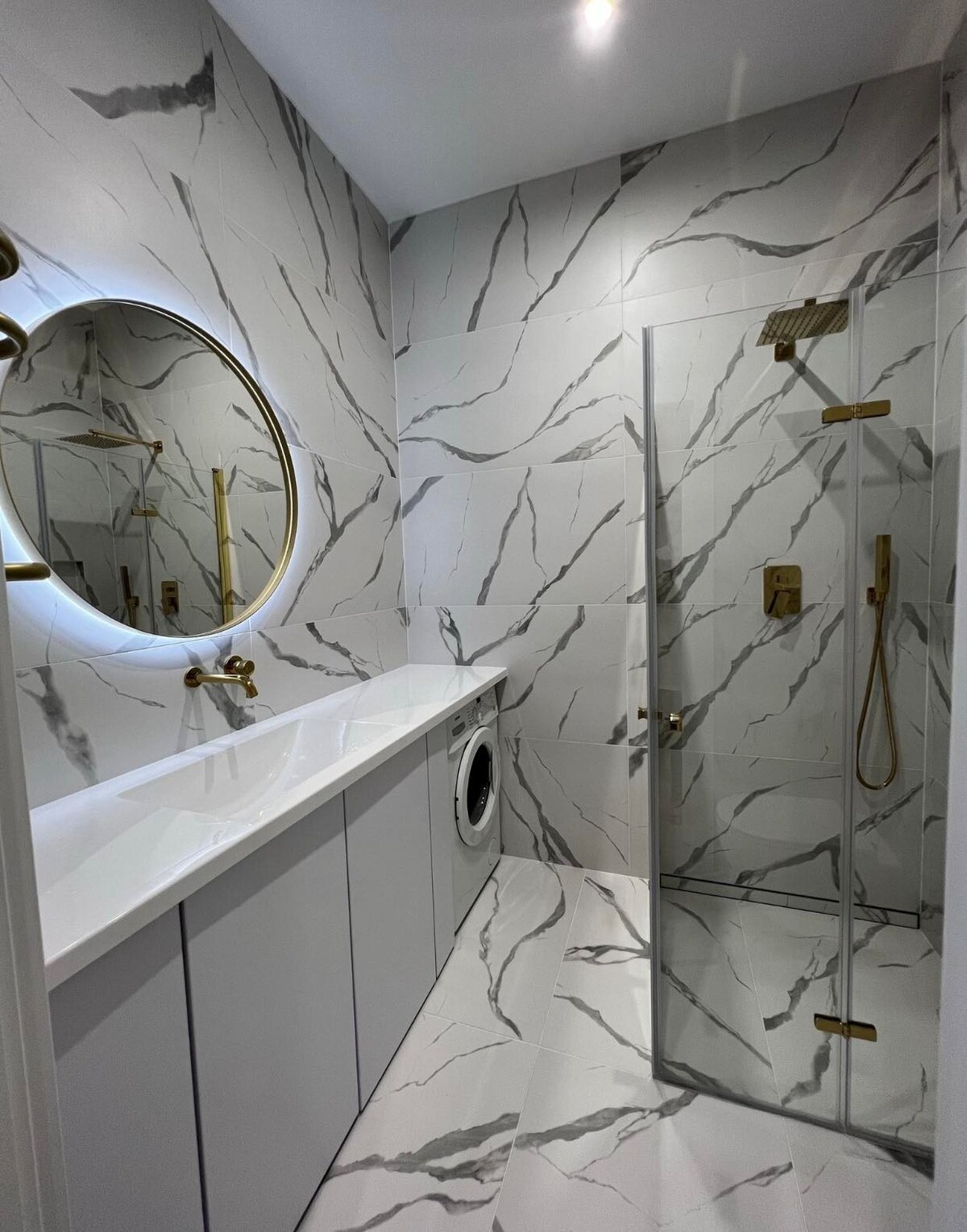 A modern bathroom features elegant marble-patterned tiles on the walls and floor. A round mirror is positioned above a sleek countertop, complemented by a washing machine integrated into the cabinetry. A glass shower with gold fixtures is visible, offering a clean and stylish bathing space.