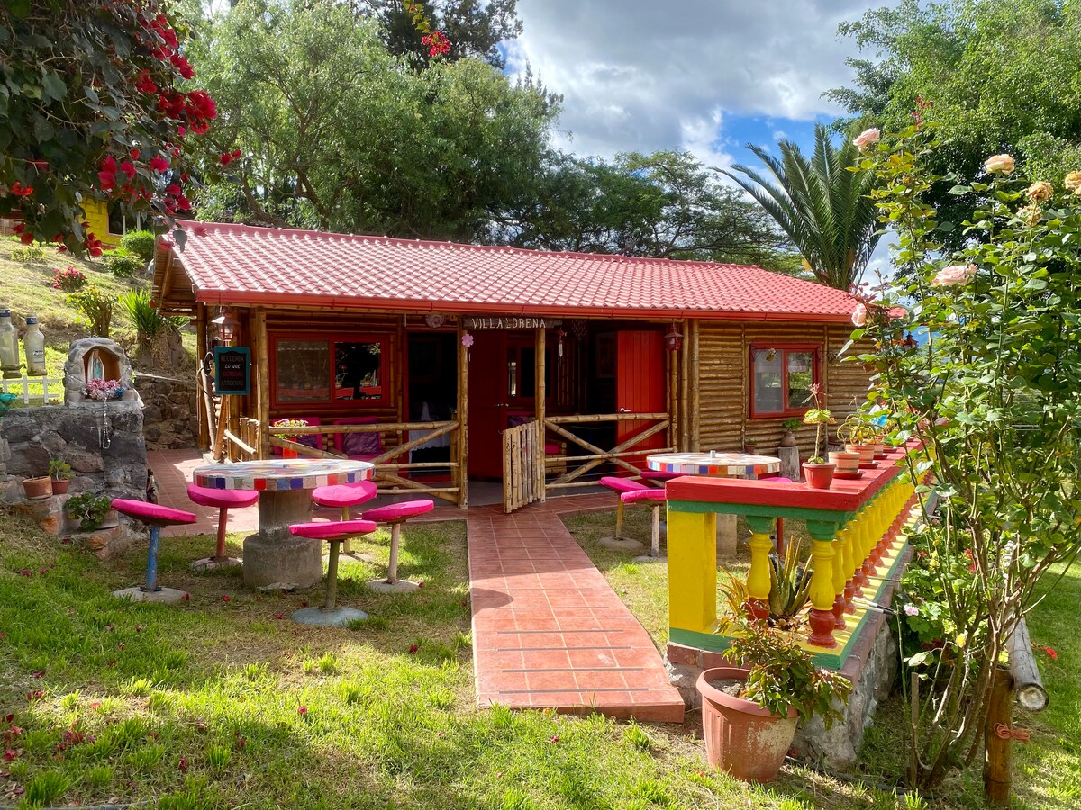A rustic wooden structure features a vibrant red roof and large windows. A pathway leads to the entrance, flanked by colorful pink tables and stone benches. Lush greenery and flowering plants surround the area, creating a welcoming outdoor space.