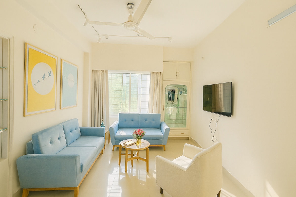 The living area features a light and airy design with soft blue sofas arranged around a wooden coffee table. A television is mounted on the wall, and natural light enters through a large window, surrounded by light-colored curtains.