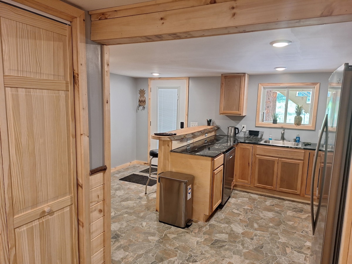 A spacious kitchen area is seen, featuring wooden cabinetry and a granite countertop. Stainless steel appliances are positioned conveniently. A large window invites natural light, while a modern bar with seating enhances the openness of the space.