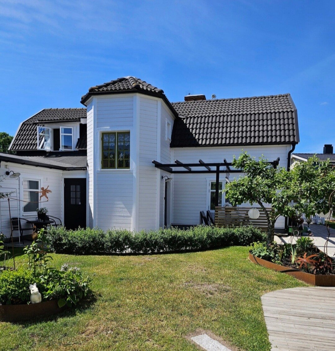 A charming white house features a unique tower element, surrounded by a well-maintained garden. Lush greenery and garden beds are visible, framing the outdoor space. Sunlight enhances the inviting appearance of the home, showcasing a blend of modern and traditional architectural elements.