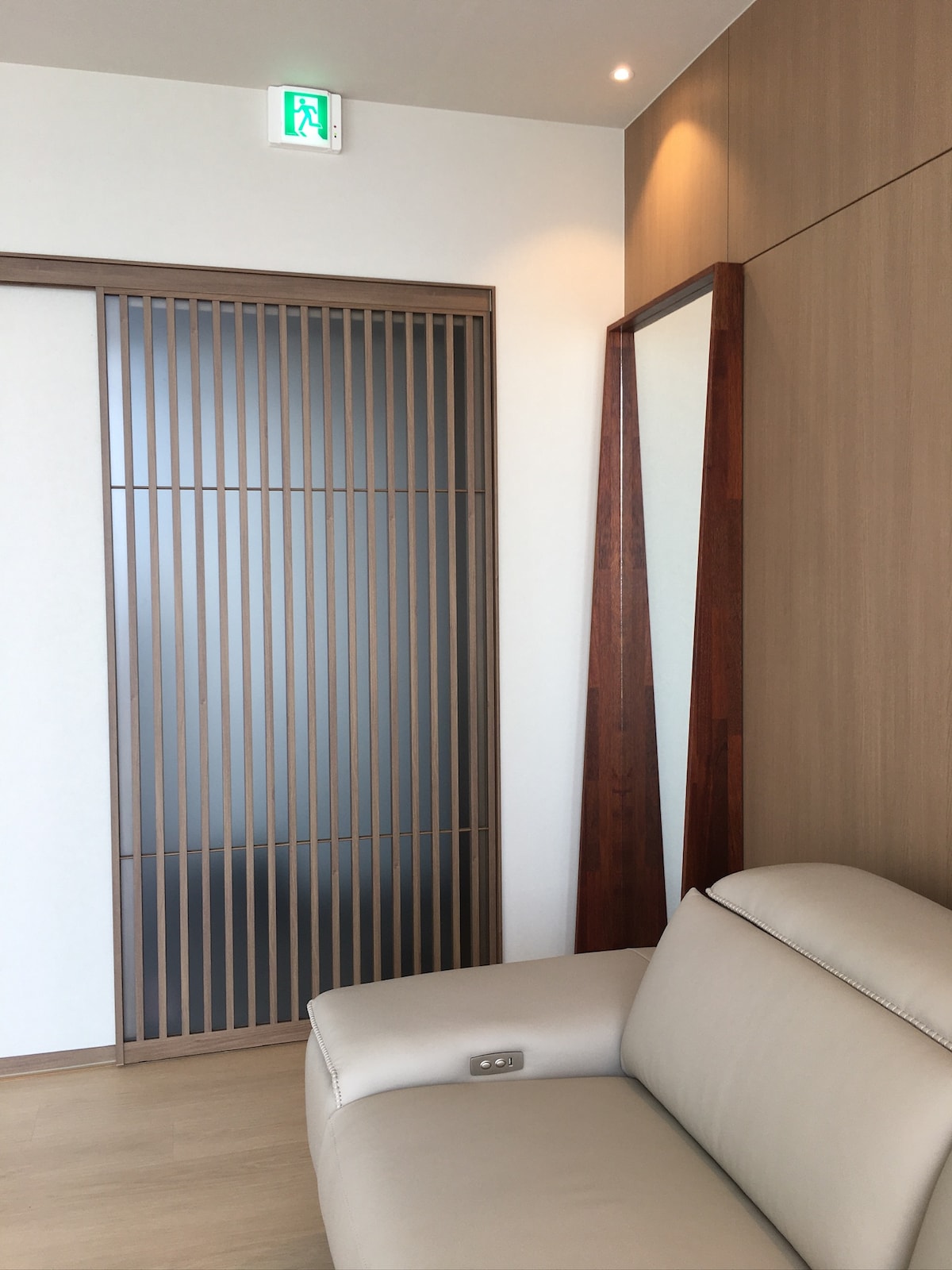 A cozy corner is presented with a light-colored sofa positioned beside a uniquely designed wooden panel. A tall mirrored wall reflects the natural light, while a sleek door featuring horizontal slats adds a modern touch to the understated decor.