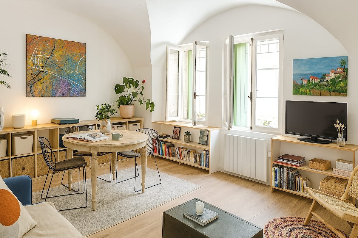 A sunlit living area is present, featuring a round table surrounded by two chairs. Shelves with books and decorative items are positioned along the walls. Two open windows allow natural light to fill the space, complemented by plants and artwork on display.