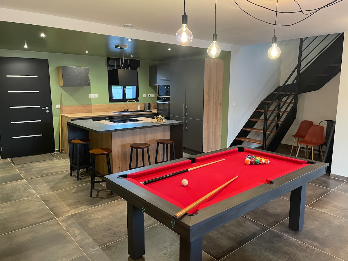 An open-plan living area features a bright red billiard table at the center, surrounded by wooden stools. The modern kitchen is visible in the background, displaying sleek cabinetry. A staircase with minimalist design leads to an upper level, with industrial-style lighting enhancing the space.