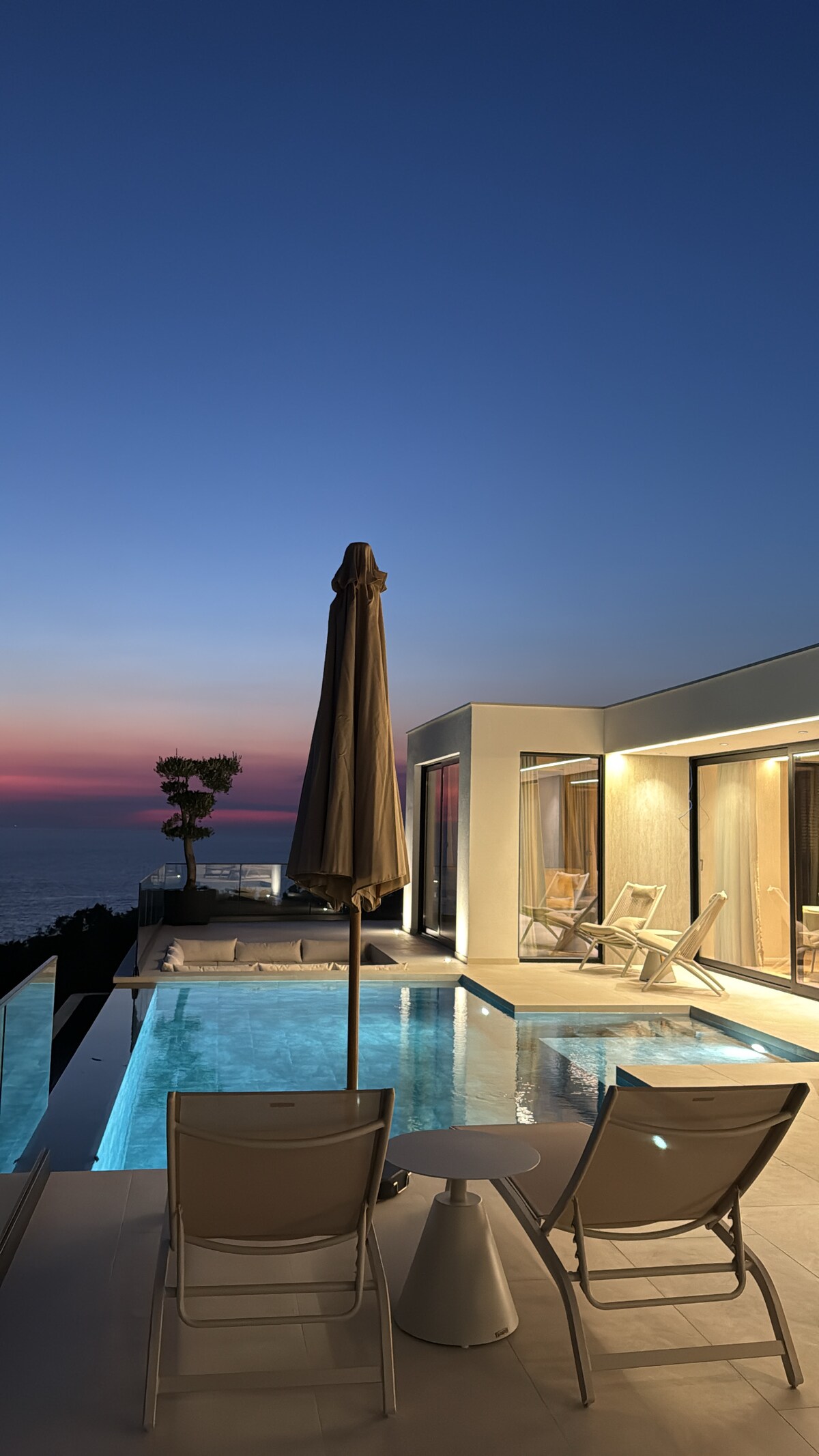 An outdoor relaxation area features two lounge chairs and a table beside a serene infinity pool. The horizon displays a gradient of colors as the sun sets over the Adriatic Sea, while a tall umbrella casts shade over the seating area.