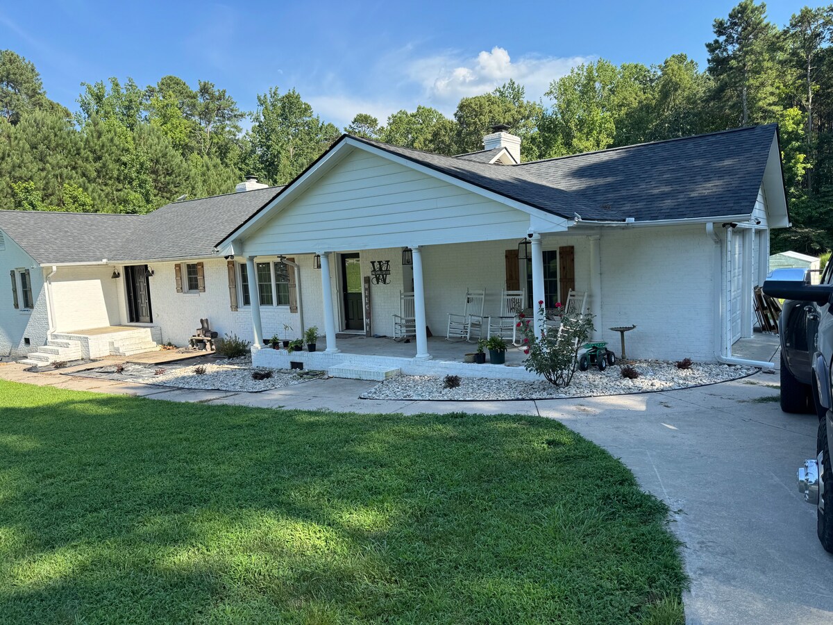 A spacious single-story home is surrounded by lush greenery and a manicured lawn. A large porch is furnished with several rocking chairs, inviting relaxation. A circular driveway leads to the entrance, enhancing accessibility for guests.
