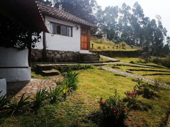 A charming cottage is situated amidst greenery, featuring a welcoming wooden door and stone steps leading to the entrance. Paths of stones traverse the lush lawn, complemented by neatly arranged flower beds and tall trees in the background, creating a serene natural setting.