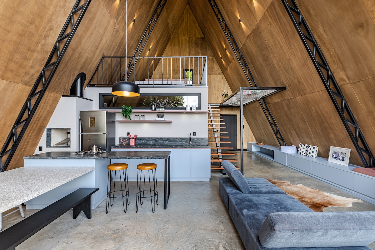 An open space features a modern kitchen with sleek cabinetry and a spacious island, complemented by two round stools. A living area is adorned with a sectional sofa and a warm-toned rug, while a staircase leads to an upper level. Natural light floods through the roof's angled structure.