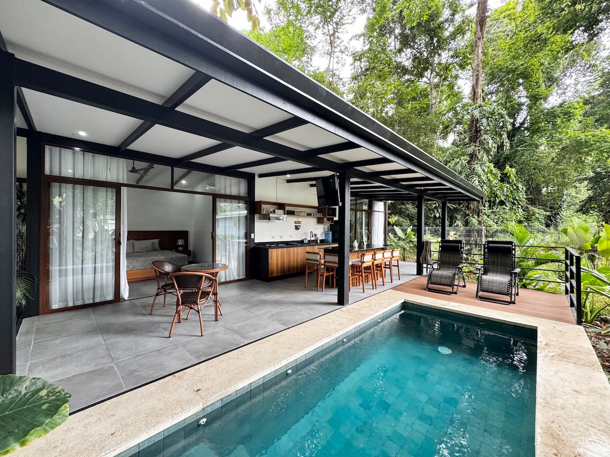 An open-concept living area flows seamlessly onto a wooden deck, where a swimming pool reflects the greenery surrounding it. The inviting space showcases comfortable seating arrangements and a dining area with a table for six. Lush vegetation provides a natural backdrop, enhancing the tranquil setting.