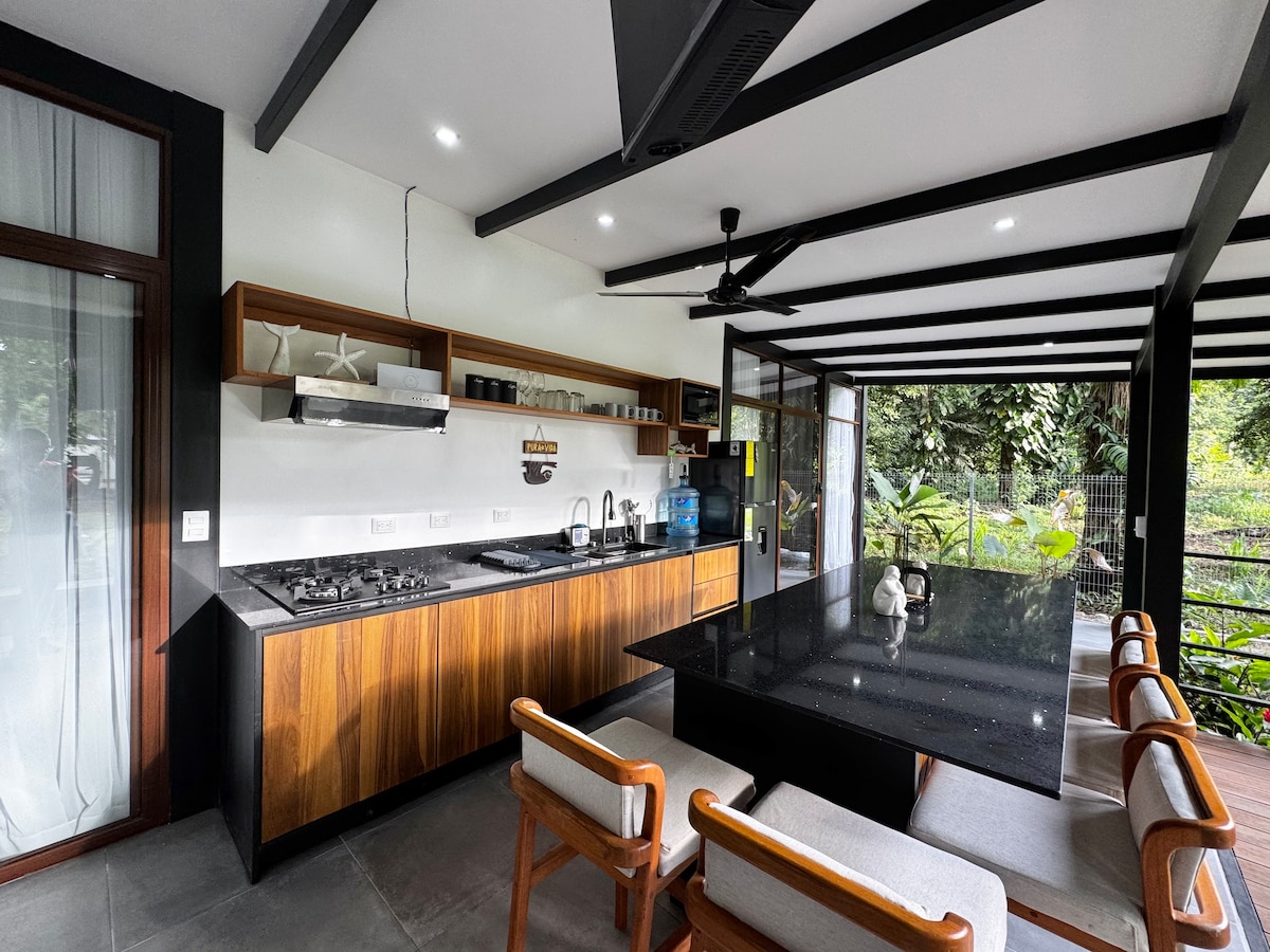 The open-kitchen area features a dark countertop complemented by wooden cabinetry. A gas stove and various utensils are visible, along with a large dining table surrounded by six chairs. Glass doors offer a view of the lush greenery outside.