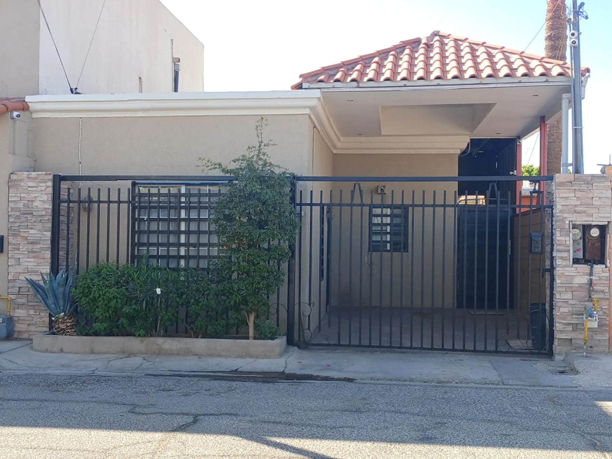 A single-story home is enclosed by a sturdy black gate and stone wall, emphasizing security and privacy. The roof is adorned with traditional red tiles. A small garden with greenery is situated near the entrance, contributing to a welcoming exterior.