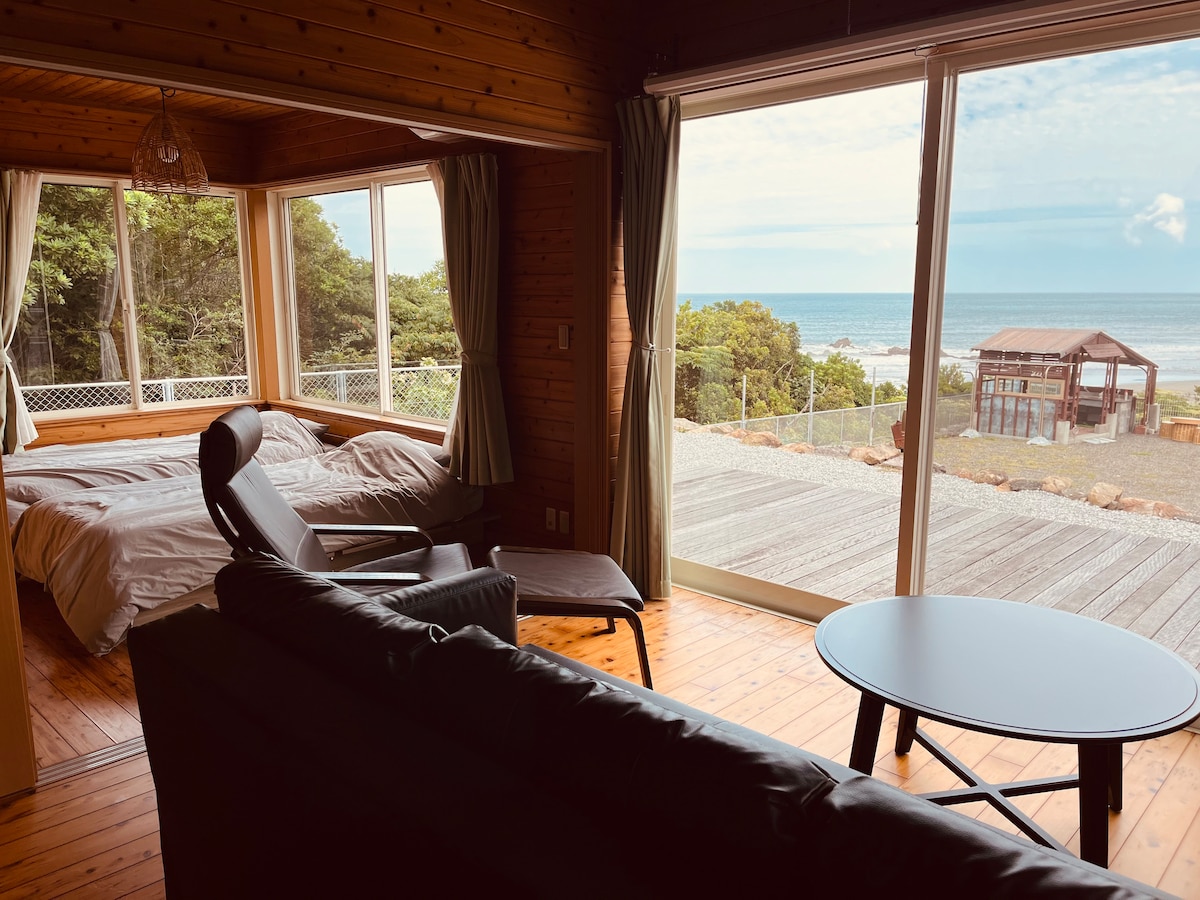 The interior features a cozy living area with a dark sofa and a round table. Large windows provide a view of the ocean and deck, while two single beds are visible in a separate area, surrounded by wooden walls.