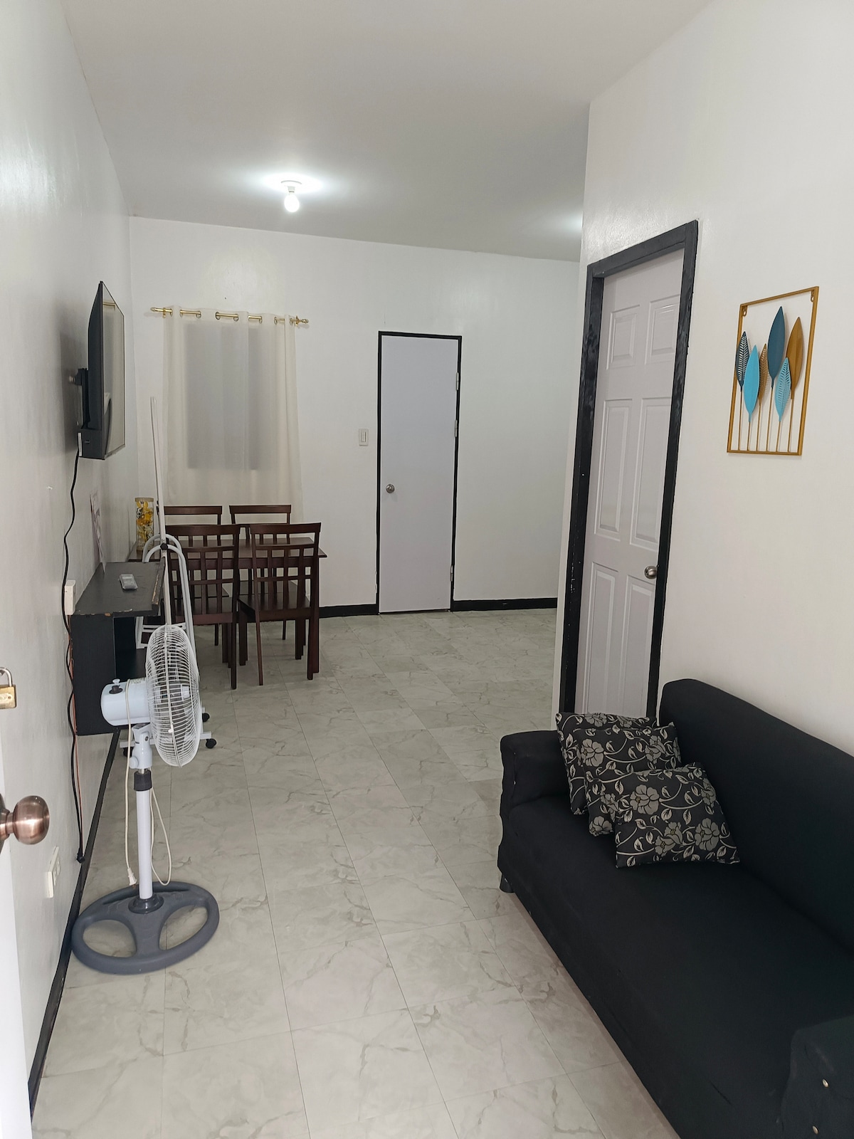 A cozy living area features a dark sofa with decorative pillows. A small dining table with chairs is positioned nearby, and a wall-mounted television is visible. The room is illuminated by overhead lighting, with a door leading to another space and a window dressed with light curtains.