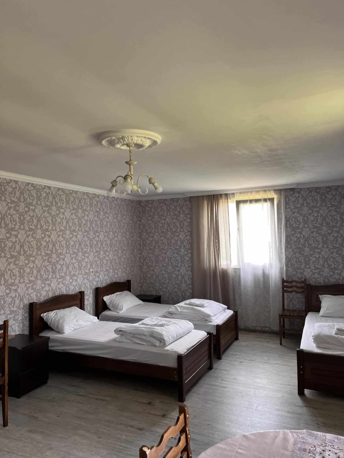 A spacious room is furnished with four wooden beds, each topped with neatly arranged linens. Light filters through a window, illuminating the soft floral wallpaper and enhancing the simple decor. A chandelier hangs from the ceiling, complementing the warm, inviting ambiance.