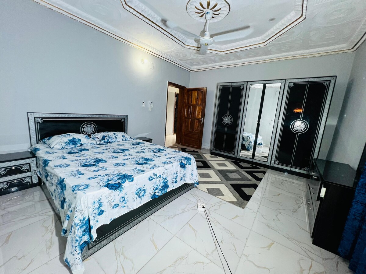 A spacious sleeping area features a floral-patterned bedspread atop a large bed. Mirrored wardrobes line one wall, reflecting light. A textured rug anchors the room, and a ceiling fan is installed above, contributing to the overall serene environment.