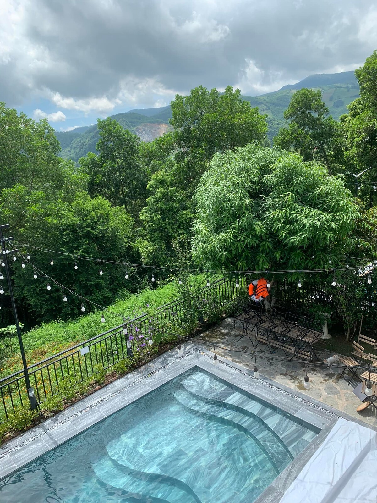 A serene outdoor pool is framed by lush greenery and mountain views in the background. String lights are suspended above the area, adding a touch of charm. The pool features a clear, inviting surface, reflecting the surrounding nature and overcast sky.