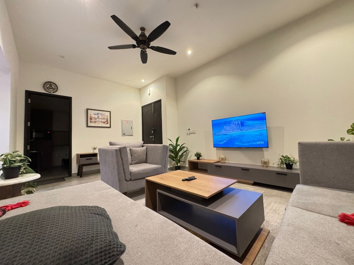 A contemporary living area features a grey sectional sofa positioned around a coffee table. A 55-inch Smart TV is mounted on the wall, and several potted plants add greenery to the space. Soft lighting creates a comfortable ambiance, while a ceiling fan offers additional airflow.