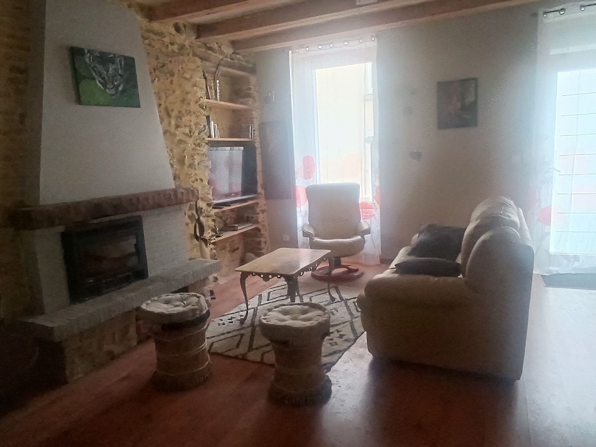 A living area features a stone fireplace, complemented by a comfortable sofa and armchair. Two round stools are present, and natural light enters through the windows, enhancing the overall ambiance of the space.