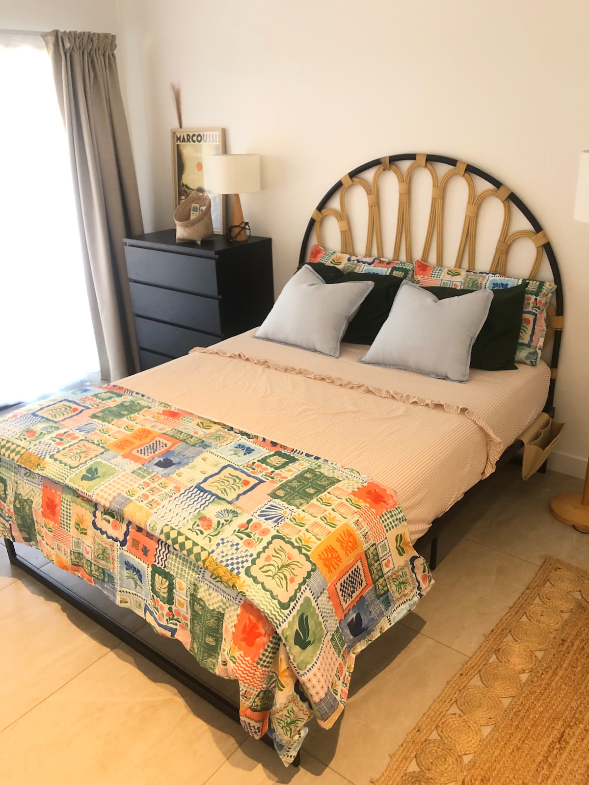 A cozy bedroom is presented with a stylish bed featuring a rounded headboard. Colorful bedding and decorative pillows brighten the space. A large window allows natural light to enter, complemented by soft curtains. Nearby, a modern dresser and a lamp add functionality and charm to the room.