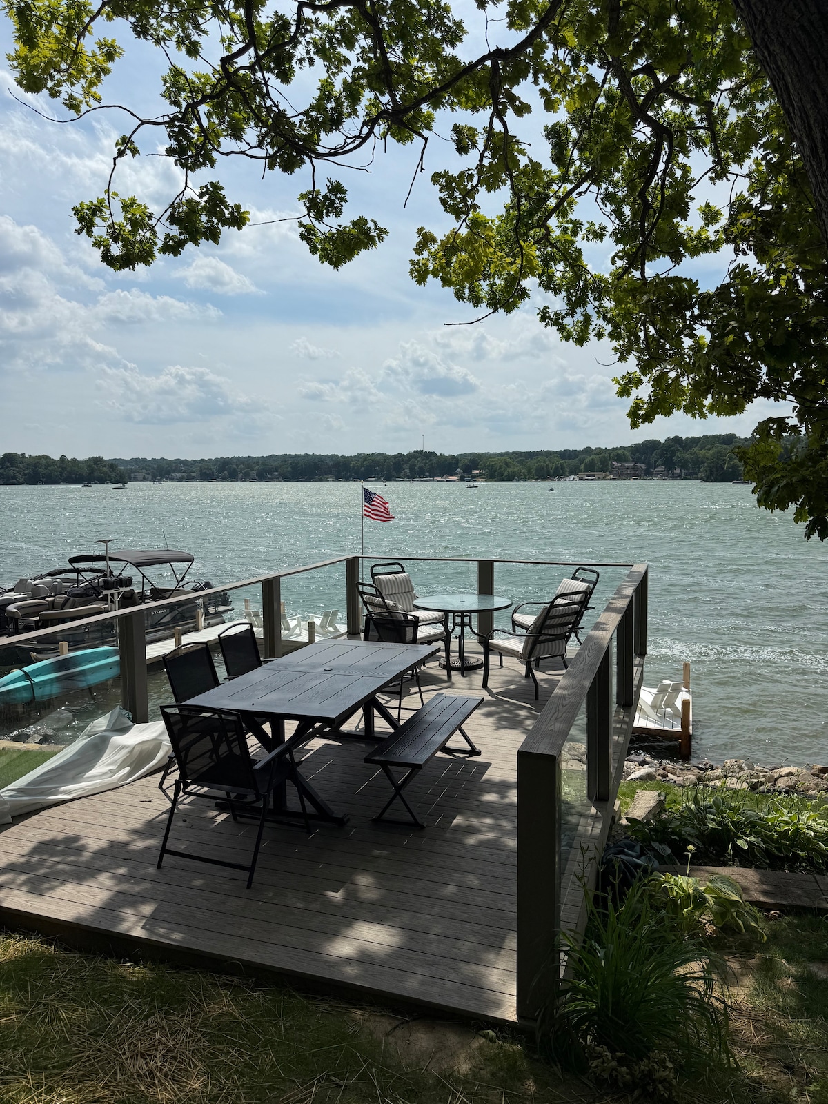 A dining area is set on a wooden deck, surrounded by glass panels. A large table with chairs faces the water, showcasing a view of the lake. An American flag gently waves nearby, framed by green leaves from a tree.