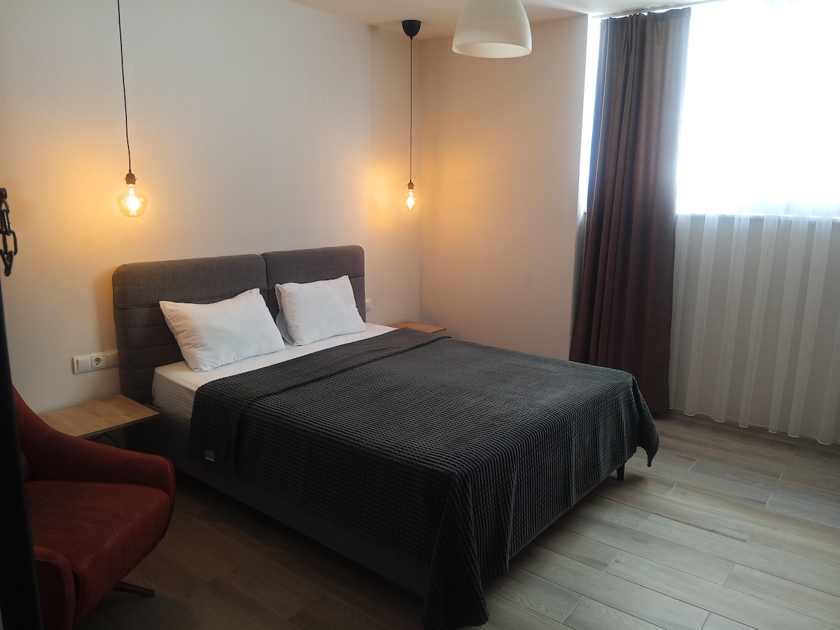 A spacious sleeping area features a comfortable bed with white linens and a textured dark blanket. Warm light is provided by hanging pendant fixtures. A modern reading chair in red adds a pop of color, while softly draped curtains frame a large window.
