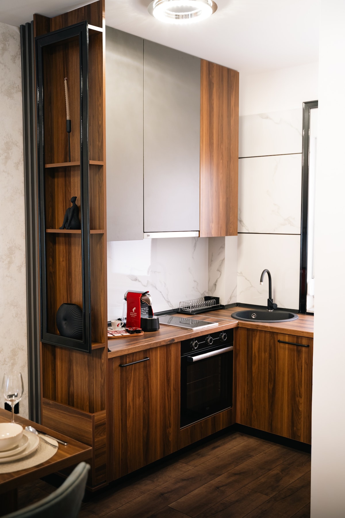 A compact kitchen area features sleek wooden cabinetry and modern appliances. A glossy black sink is set against a marble-patterned backsplash. An oven is integrated into the cabinetry, with additional counter space for meal preparation, and a small decorative item is displayed on a shelf.