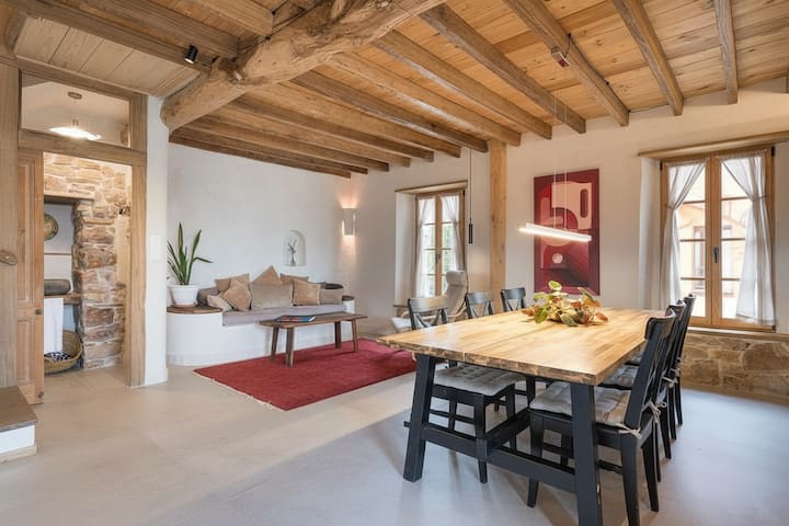 Warm, open-plan living room with exposed chestnut beams, a large wooden dining table, built-in sofa with neutral cushions, soft natural light through wooden-framed windows, stone details, and minimalist décor for a cozy, eco-luxury feel.