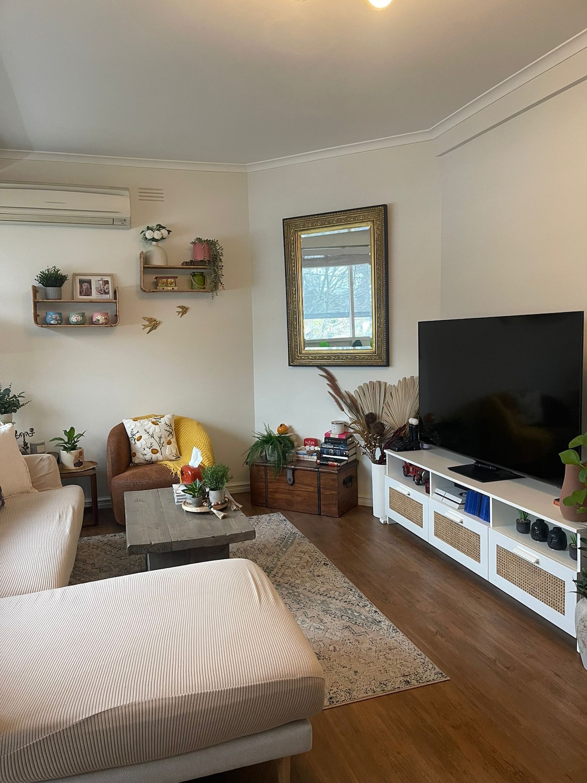 A cozy living room is presented, featuring a light-colored sectional sofa and a patterned area rug. A large television is mounted on a white media console. Decorative plants and a mirror accentuate the space, complemented by a small coffee table adorned with various items.