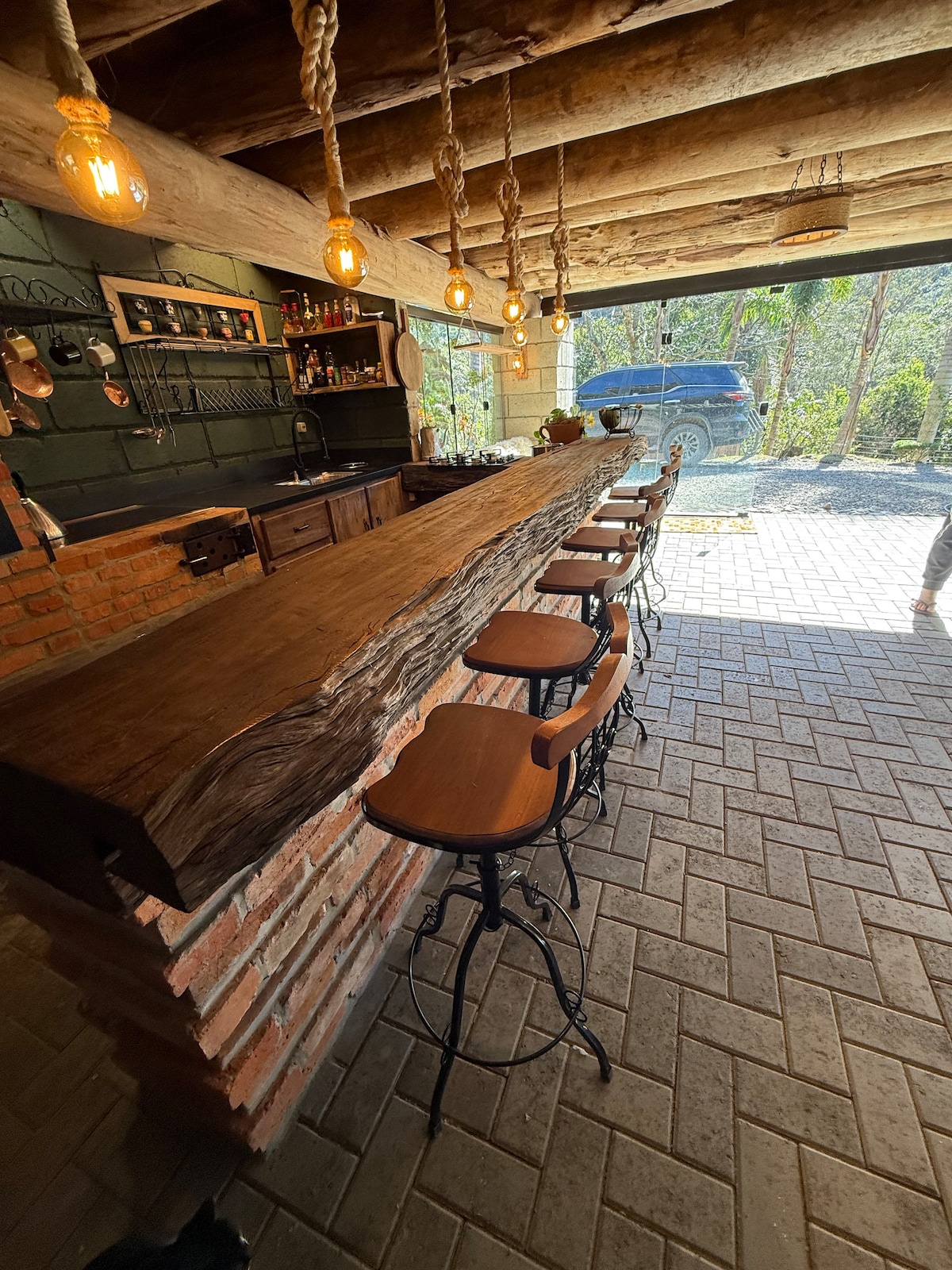 A rustic kitchen bar features a long wooden countertop supported by brick pillars. Several high-back stools are arranged along one side, with warm pendant lights hanging above. Natural light floods the space from large windows, creating a connection with the surrounding landscape.