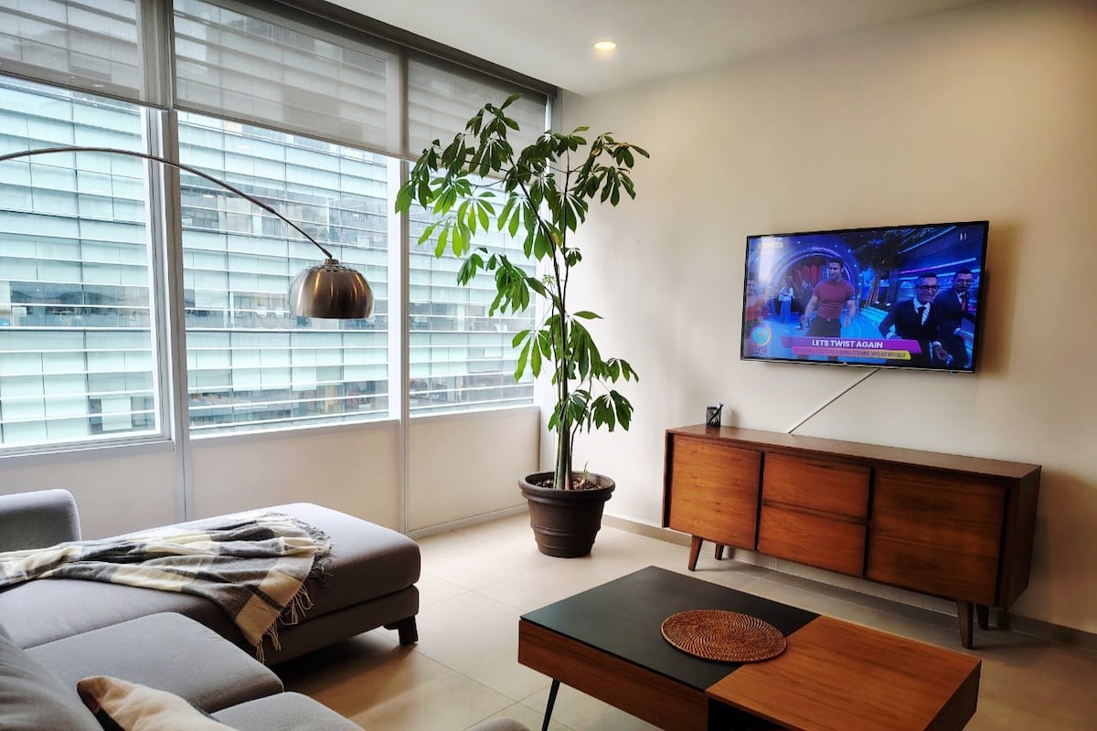 A spacious living area features a comfortable sectional sofa, complemented by a floor lamp. A large potted plant adds a touch of greenery. A television mounted on the wall displays content, while a sleek wooden coffee table enhances the modern design. Large windows allow natural light to fill the room.