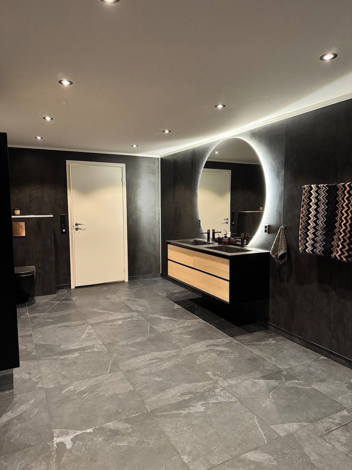 A spacious bathroom features dark slate flooring and contemporary lighting. A circular mirror above a wooden vanity reflects the modern design. The room includes a towel rack, a wall-mounted shelf, and a discreet toilet area, creating a cohesive and functional layout.