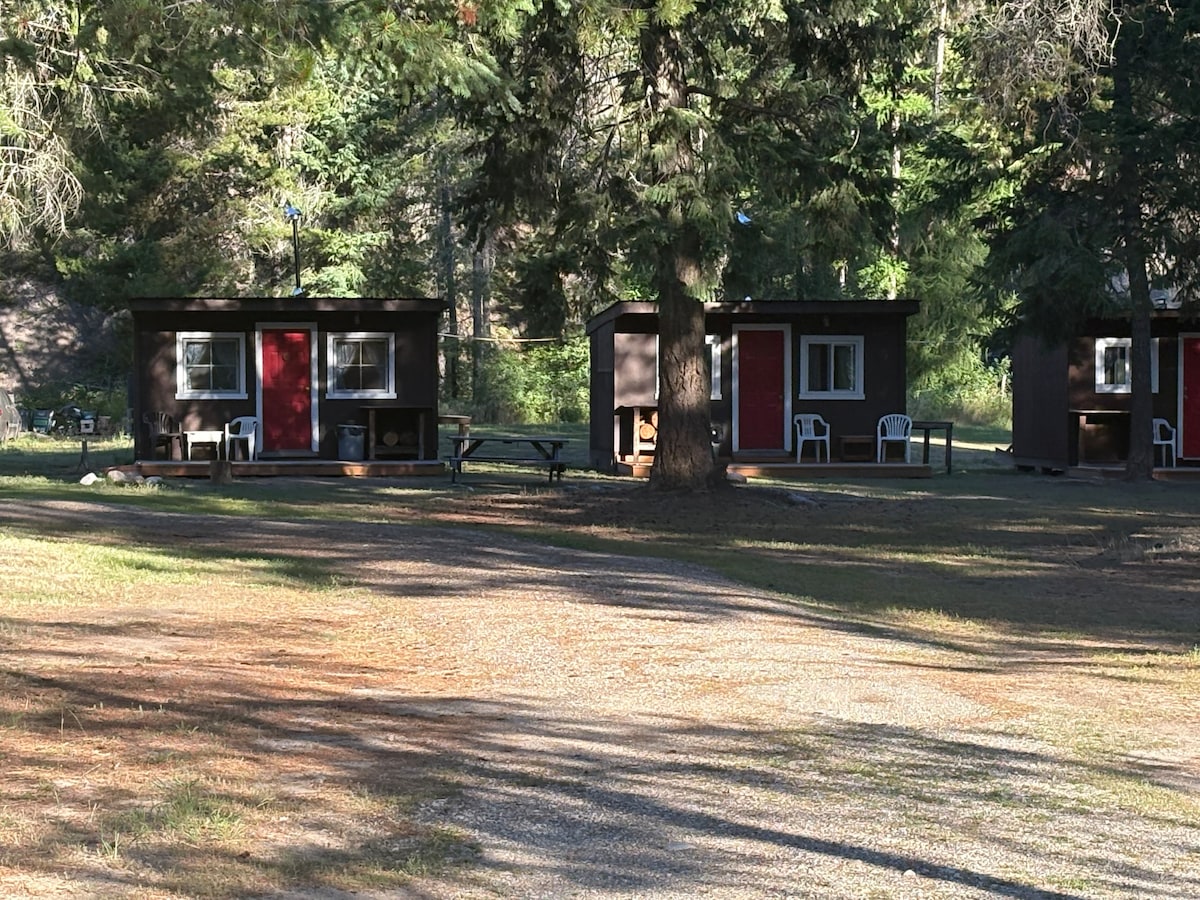 Two cabin-like structures are visible, each featuring a red door and situated in a wooded area. A gravel path leads to the cabins, and outdoor seating options are provided nearby. Tall trees surround the space, offering a natural and serene environment.
