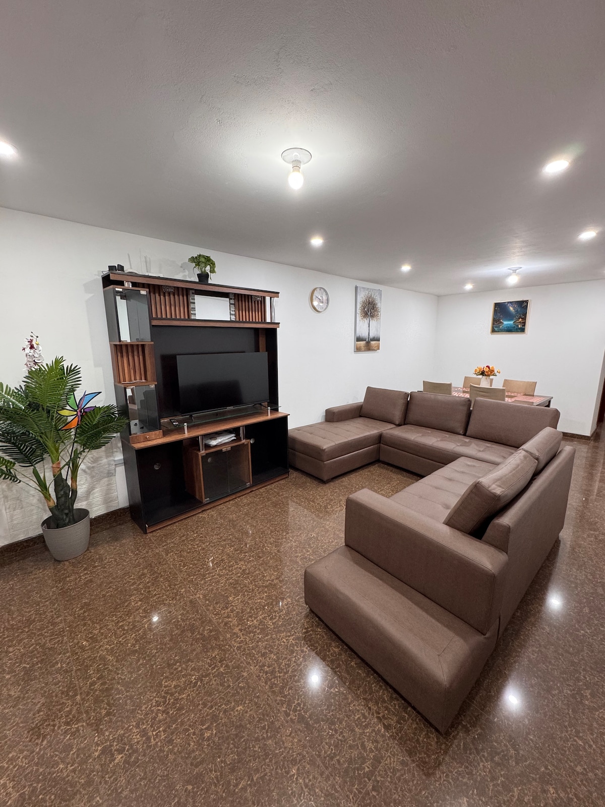 A spacious living room features a modern sectional sofa in a neutral tone, complemented by a stylish entertainment unit with a large TV. Subtle decorative elements, including a potted plant, add a touch of greenery, while soft lighting enhances the inviting atmosphere.