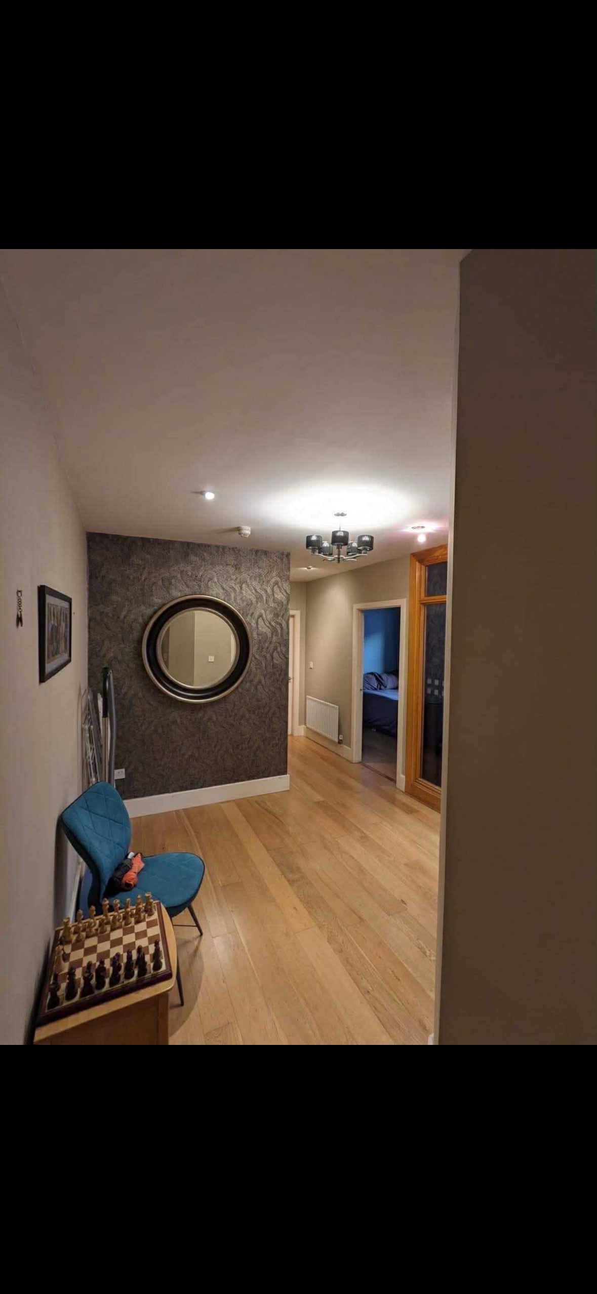 A hallway features a round mirror mounted on a wallpapered wall with a subtle pattern. A blue chair is positioned next to a small chessboard on a wooden bench. Soft lighting is provided by ceiling fixtures, illuminating the space.