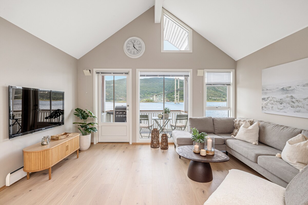 A light-filled living area features large windows that overlook the water and mountains. Comfortable seating is arranged around a round coffee table, with a wall-mounted television providing entertainment. A subtle coastal decor element is visible, complemented by a plant in the corner.