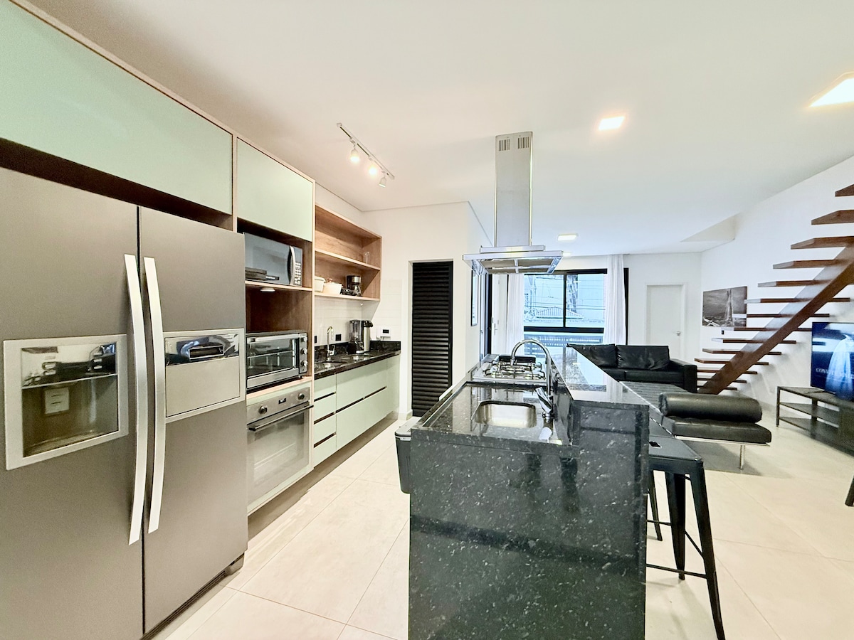 A modern kitchen features sleek cabinetry and high-end appliances, including a side-by-side refrigerator and a gas cooktop. An island with seating provides a functional workspace. The room flows openly into a living area, illuminated by natural light from adjacent windows.